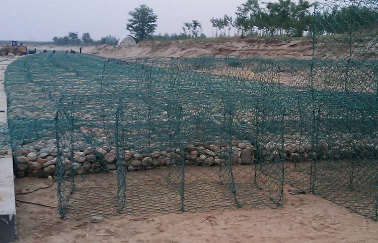 Green PVC coated gabion boxes in the river bank construction site. Construction workers are in the distance, nearby, there are a few empty boxes just installed, not filled stone.